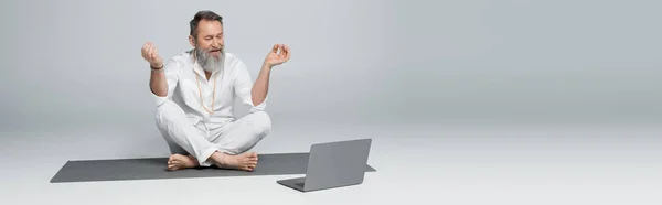 Senior yoga coach showing chin mudra near laptop while sitting in easy pose on grey, banner — Stock Photo