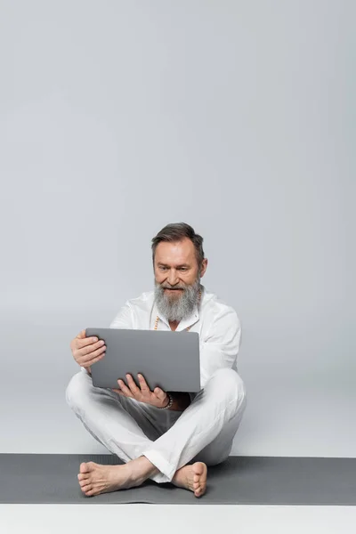 Senior yoga coach with laptop sitting on yoga mat in easy pose on grey — Stock Photo