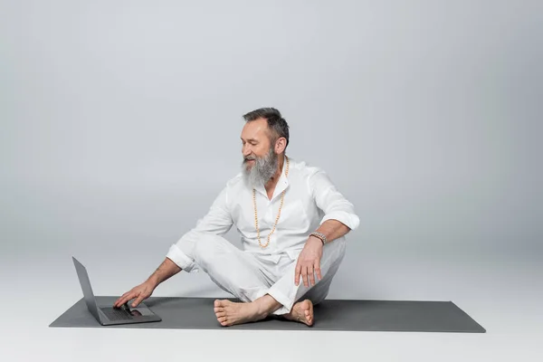 Smiling spiritual mentor sitting on yoga mat in easy pose near laptop on grey — Stock Photo