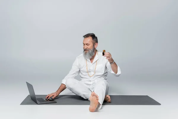 Gurú sanador descalzo sentado cerca de la computadora portátil y sosteniendo la botella de aceite perfumado en gris - foto de stock