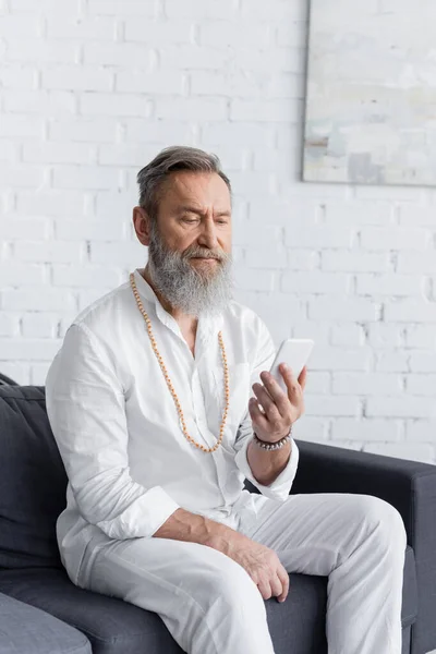 Bearded guru man in white shirt and beads sitting on couch with mobile phone — Stock Photo