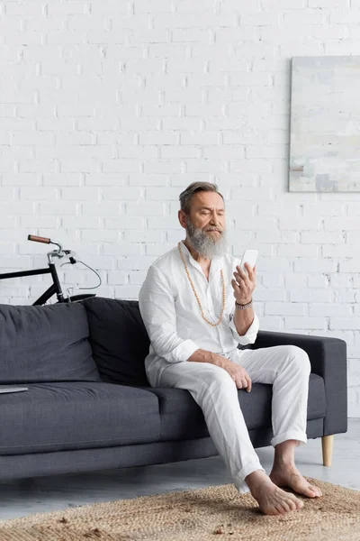 Anziano guru uomo a piedi nudi con telefono cellulare seduto sul divano a casa — Stock Photo