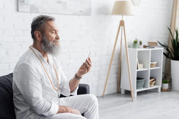 Bearded guru man in white clothes sitting on sofa and looking at mobile phone — Stock Photo