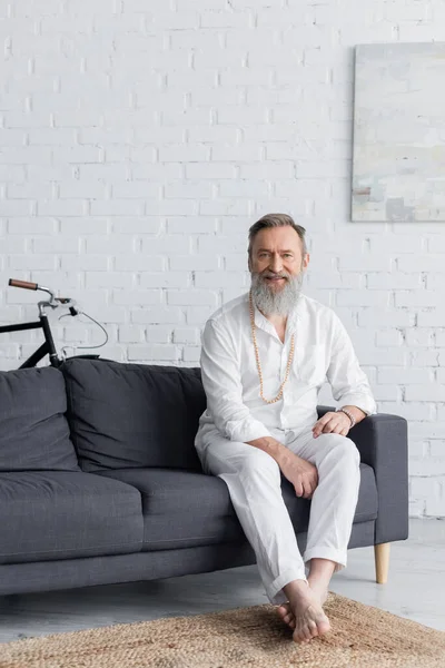 Maître gourou senior en vêtements blancs assis sur le canapé à la maison et souriant à la caméra — Photo de stock