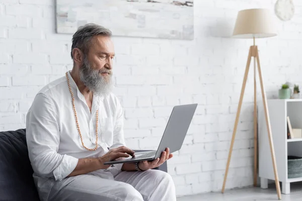 Barbuto allenatore spirituale in abiti bianchi seduto sul divano e utilizzando il computer portatile — Stock Photo