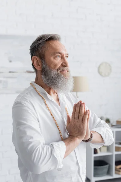 Hombre enfocado mostrando anjali mudra y mirando hacia otro lado mientras medita en casa - foto de stock