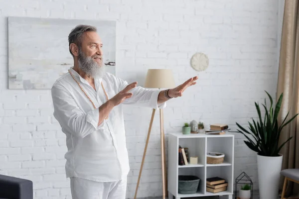 Gurú barbudo hombre de camisa blanca meditando con las manos extendidas en casa - foto de stock