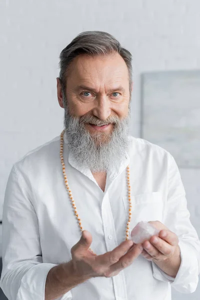 Pleased senior master guru holding selenite crystal and smiling at camera — Stock Photo