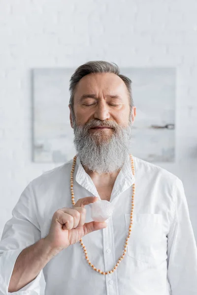 Homem guru barbudo meditando com olhos fechados e pedra selenita em casa — Fotografia de Stock