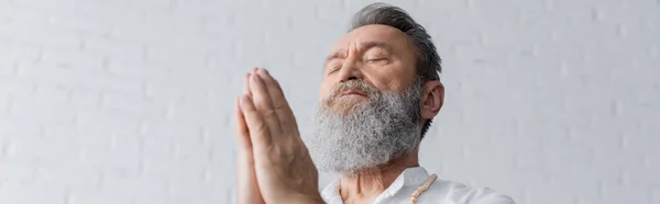 Vue à angle bas du gourou homme avec barbe grise méditant avec les yeux fermés et les mains priantes, bannière — Photo de stock