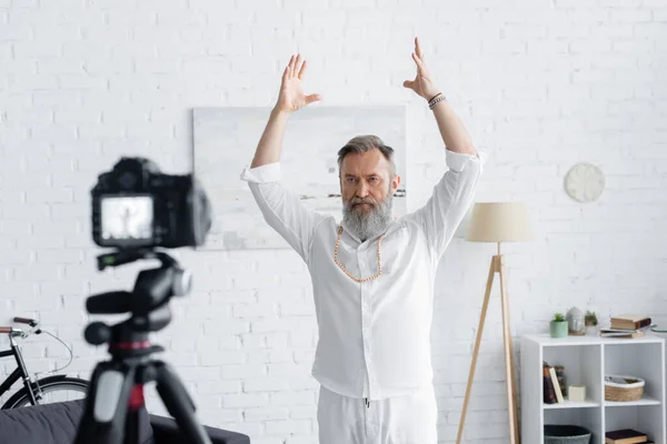 Bearded spiritual mentor meditating with raised hands near blurred digital camera at home — Stock Photo