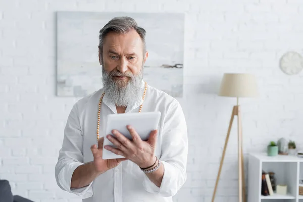 Mentor espiritual sênior em camisa branca e pulseiras de contas usando tablet digital — Fotografia de Stock