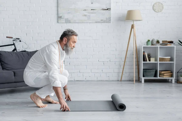 Vista lateral del hombre mayor en ropa blanca desenrollando estera de yoga en casa - foto de stock