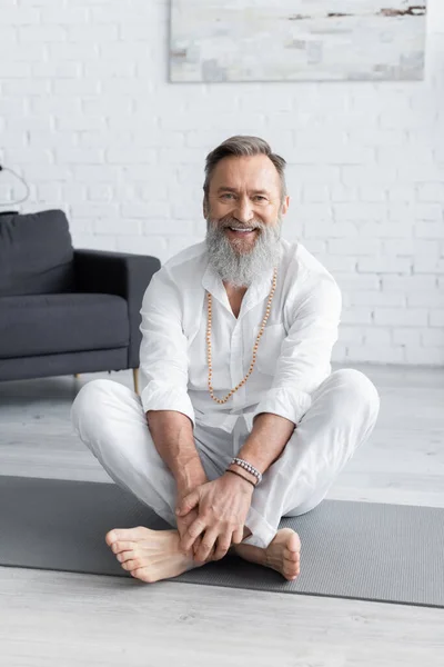 Счастливый тренер по йоге в браслетах с бисером, сидящий в легкой позе и смотрящий в камеру — стоковое фото