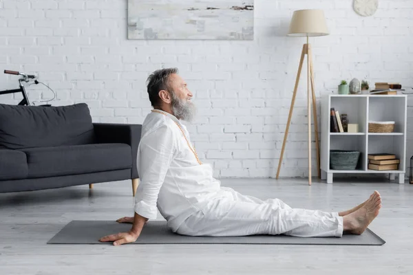 Side view of bearded guru man sitting on yoga mat while meditating at home — Stock Photo