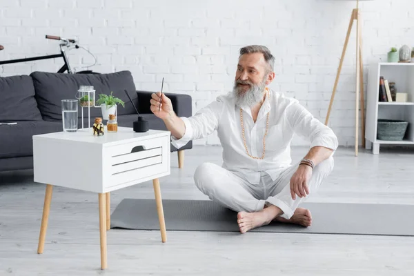 Guru sorridente homem segurando vara aroma enquanto sentado em pose fácil perto de mesa de cabeceira com óleos essenciais — Fotografia de Stock