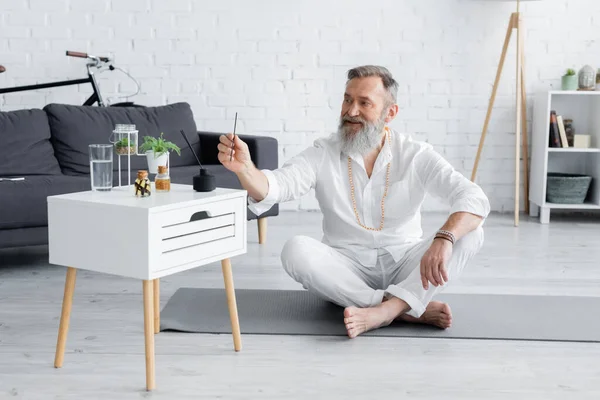 Sonriente gurú hombre celebración aroma palo mientras sentado en fácil pose cerca de mesita de noche con aceites esenciales - foto de stock