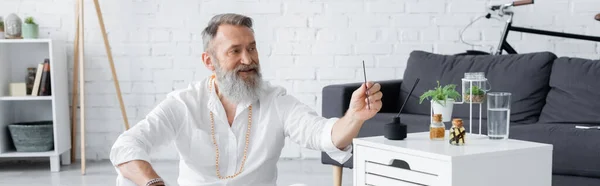 Guru de cura barbudo segurando vara de aroma perto de mesa de cabeceira com óleos essenciais, banner — Fotografia de Stock