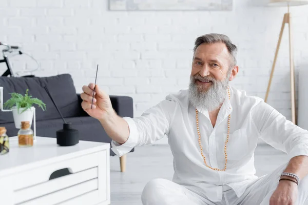 Happy ayurveda guru holding aroma stick near blurred nightstand with flavored oils — Stock Photo