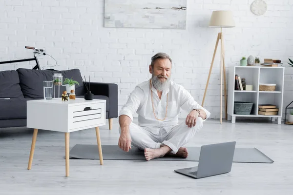 Spiritual healing master in easy pose looking at laptop near nightstand with essential oils and aroma sticks — Stock Photo
