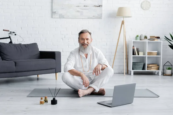 Treinador de ioga sorridente sentado em pose fácil perto de óleos aromatizados e laptop — Fotografia de Stock