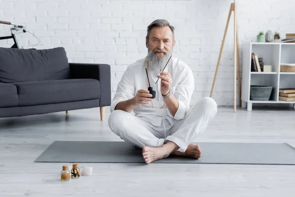 Gurú curativo barbudo sentado en pose fácil con palos de aroma cerca de botellas con aceites aromatizados - foto de stock