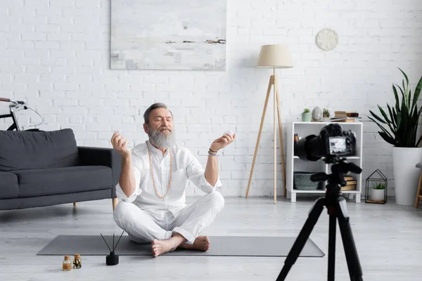 Guru treinador sênior meditando com pedras de selenita perto de óleos essenciais e câmera digital — Fotografia de Stock