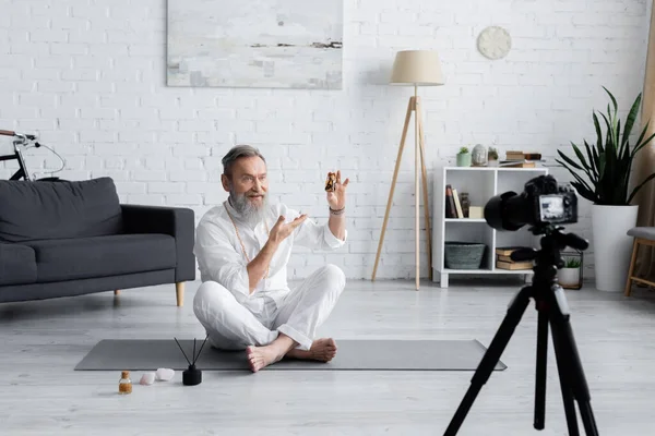 Senior-Heilcoach zeigt auf Flasche mit ätherischem Öl, während er in lockerer Pose in der Nähe der Digitalkamera sitzt — Stockfoto