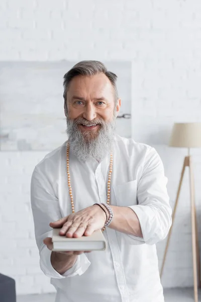 Glücklicher spiritueller Trainer, der in die Kamera blickt, während er Buch hält — Stockfoto