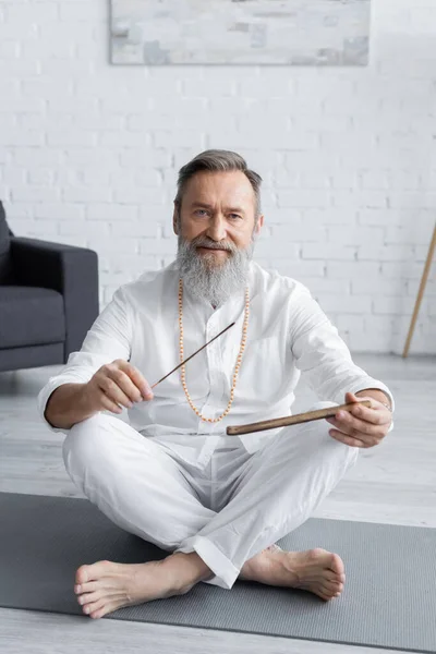 Bearded yoga teacher holding aroma sticks while sitting in easy pose and looking at camera — Stock Photo