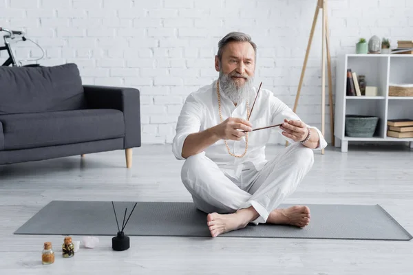 Guru sênior homem com varas de aroma sentado em pose fácil perto de óleos aromatizados — Fotografia de Stock
