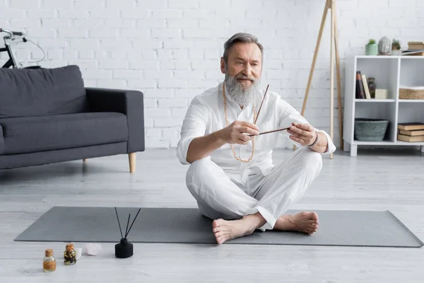 Hombre gurú senior con palos de aroma sentado en pose fácil cerca de aceites aromatizados - foto de stock