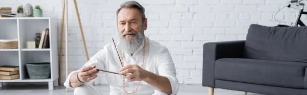 Hombre gurú senior en camisa blanca sentado cerca del sofá y sosteniendo palo de aroma, pancarta - foto de stock