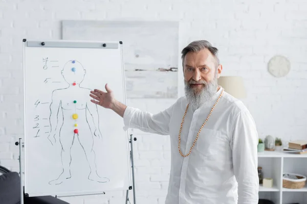 Entrenador ayurveda senior señalando el rotafolio con chakras humanos esquema y mirando a la cámara - foto de stock