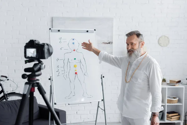 Senior ayurveda coach pointing at human chakras system on flip chart in front of digital camera — Stock Photo