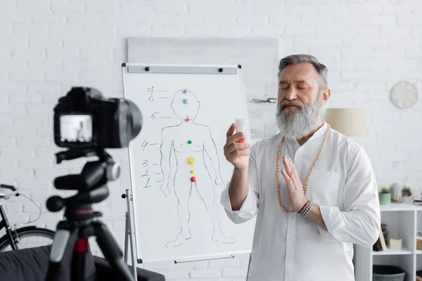 Guru espiritual meditando com pedra selenita perto da câmera digital e flip chart com esquema de chakras humanos — Fotografia de Stock