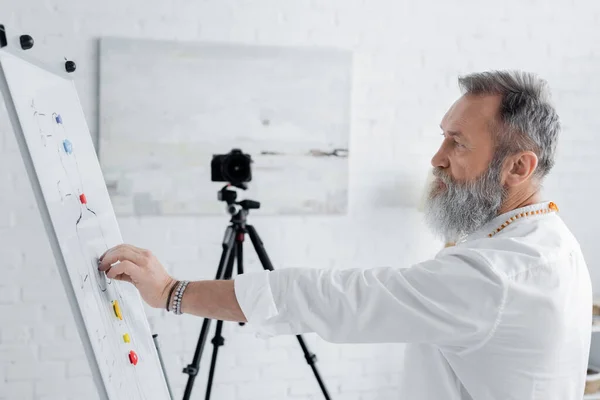 Side view of bearded healing guru near flip chart with human chakras scheme and digital camera — Stock Photo