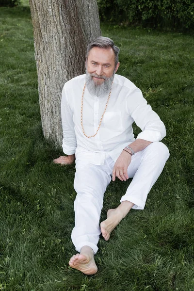 Toute la longueur du coach de yoga barbu assis sur la pelouse verte et souriant à la caméra — Photo de stock