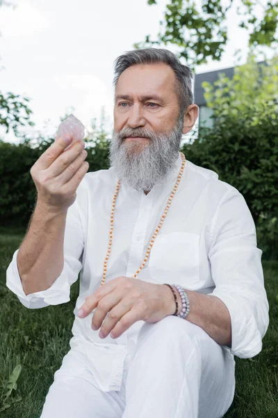 Senior master guru in white shirt and beads looking at selenite crystal outdoors — Stock Photo