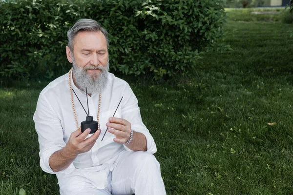 Gurú barbudo curativo sosteniendo difusor y palos de aroma mientras está sentado en el césped en el jardín - foto de stock