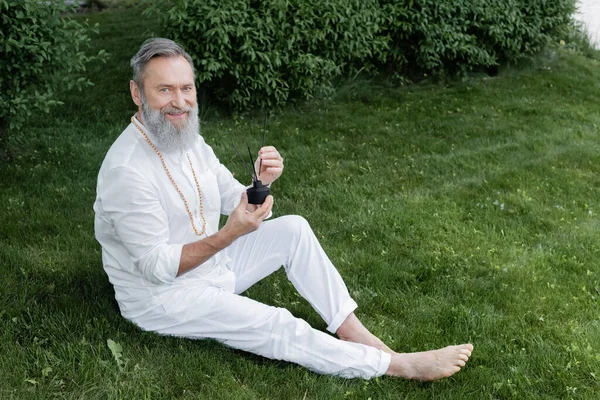 Happy senior yoga coach sitting with diffuser and aroma sticks on green lawn — Stock Photo