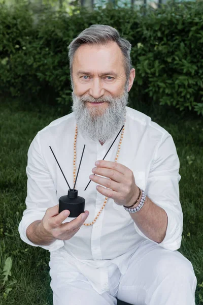 Guru mestre barbudo com varas de aroma e difusor sorrindo para a câmera ao ar livre — Fotografia de Stock