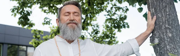 Guru sênior homem com olhos fechados meditando perto de tronco de árvore, banner — Fotografia de Stock