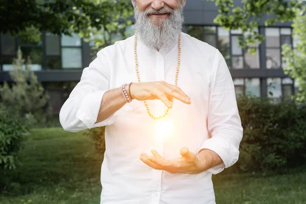 Abgeschnittene Ansicht eines lächelnden Guru-Mannes, der in der Nähe einer strahlenden Aura im Park meditiert — Stockfoto