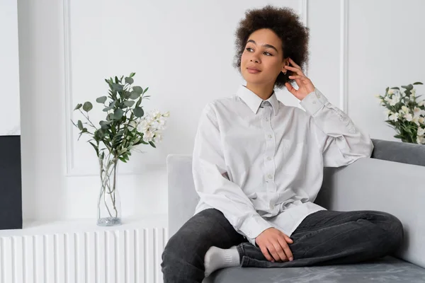 Dreamy african american woman sitting on grey velvet sofa near vase with green leaves — Stock Photo