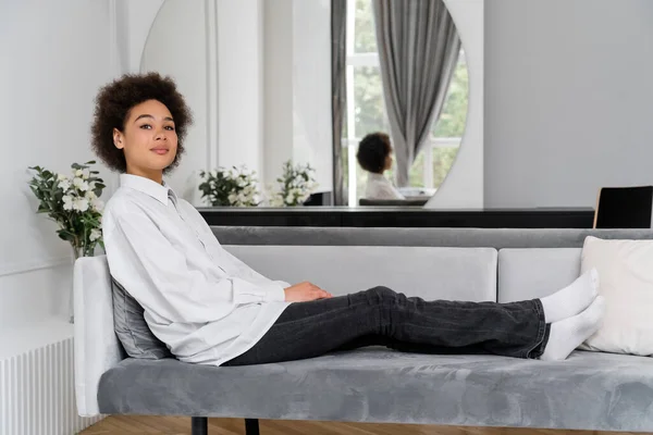 Curly african american woman resting on grey velvet sofa near vase with green leaves — Stock Photo