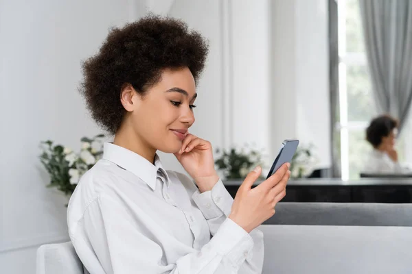 Gioiosa donna afro-americana sorridente mentre usa lo smartphone a casa — Foto stock