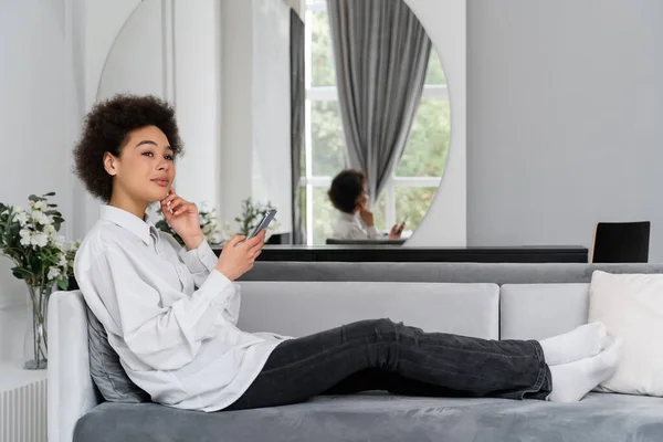 Verträumte afrikanisch-amerikanische Frau hält Smartphone in der Hand, während sie sich auf Samtsofa im modernen Wohnzimmer ausruht — Stockfoto