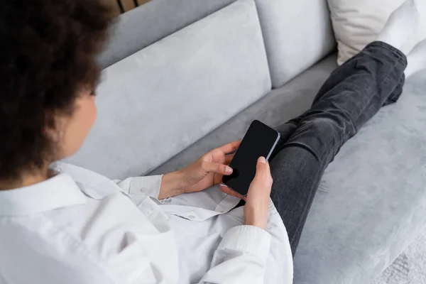 Vue grand angle de femme afro-américaine bouclée tenant smartphone avec écran vide tout en étant assis sur le canapé — Photo de stock