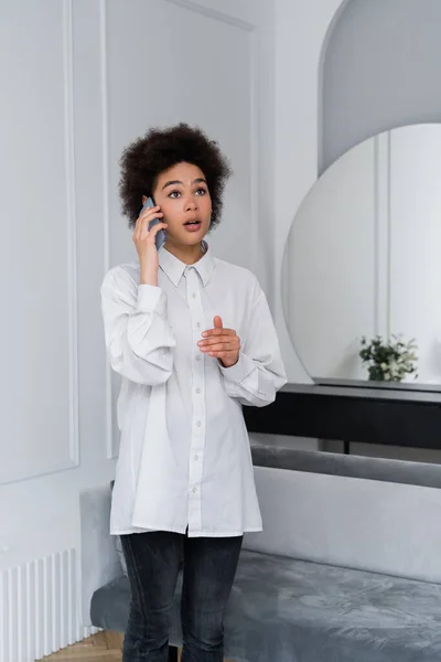 Femme afro-américaine choquée parlant sur téléphone portable dans le salon moderne — Photo de stock